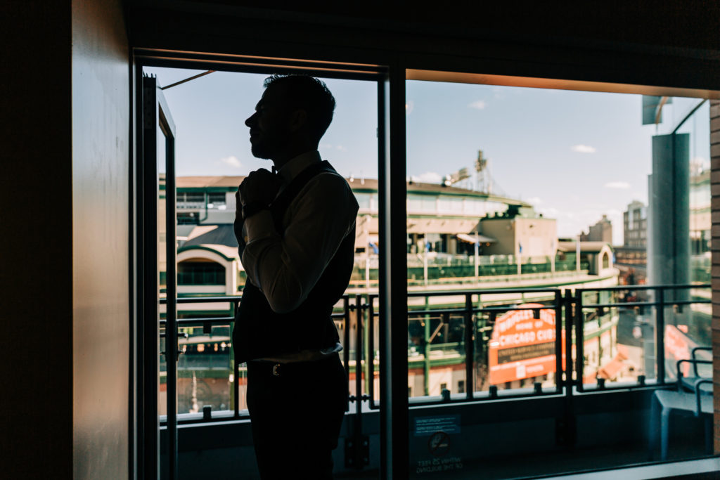 The groom ties his tie in front of a mirror, with Wrigley Field visible through the balcony windows behind him. 