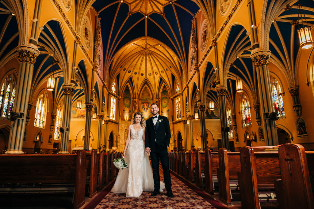 The bride and groom pose for a portrait at St. Alphonsus church, standing in the middle of the aisle and smiling at the camera.