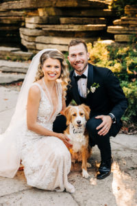 The bride and groom crouch down for a portrait with their dog. She is small in size, with long fur that is brown on her face and body, and white on her nose, chest, and paws. She is wearing a white bandana around her neck that says 'I do too.'