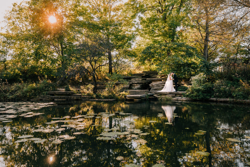 The bride and groom are seen kissing in the distance in a portrait taken at Caldwell Lily Pool in Lincoln Park. The reflection of the couple and the trees are seen among the lily pads in the pond.