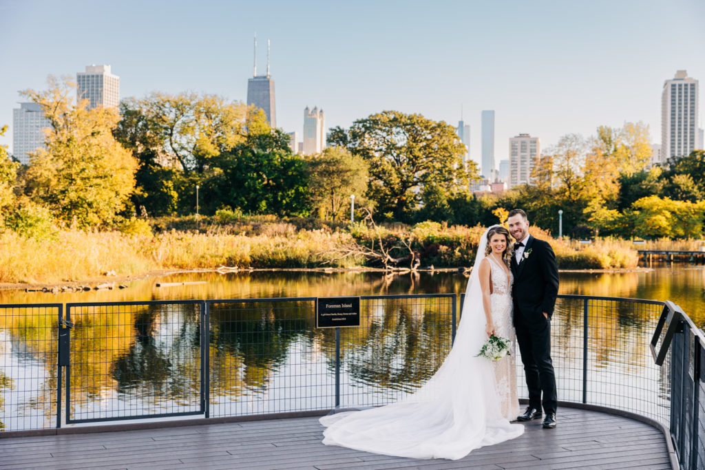 The bride and groom pose on a bridge in a park, with fall trees and the Chicago skyline behind them. 