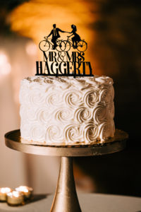 A single-tiered wedding cake sits on a tall gold cake stand. It is covered with swirls of white frosting, and has a thin black topper featuring two figures riding bikes, with the words 'Mr. and Mrs. Haggerty.'