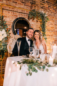 The bride and the groom are smiling and seated at a sweetheart table set for dinner, with a modern gold floral arch behind them featuring white roses, baby's breath, and eucalyptus leaves.