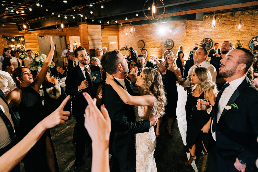 The bride and groom dance closely together on the dance floor, with their friends standing around them exuberantly singing along to their first dance song.