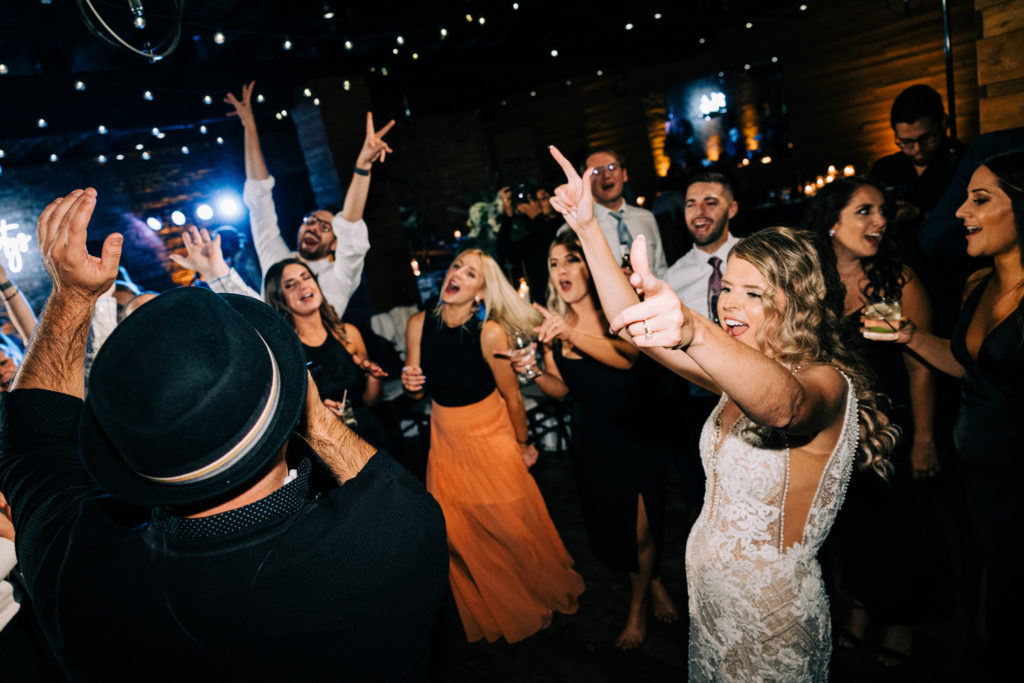 The bride dances in the middle of the dance floor with her arms in the air, surrounded by guests also dancing. They are facing the musician who is singing to the crowd.