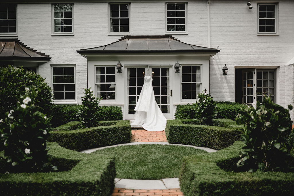 The bride's wedding dress hangs off of the French doors in the garden, framed by hedges and topiaries